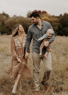 a man and woman holding hands while walking through a field with a baby in their arms