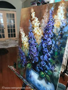 an oil painting of purple flowers in a blue vase on a easel next to a window