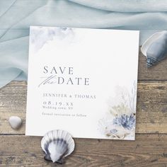 an ocean themed save the date card and seashells on a wooden table with blue fabric