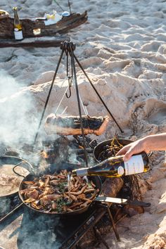 a person is cooking food on the beach with a wine bottle in their hand and another person holding a camera