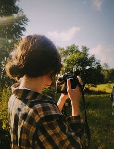 a woman holding a camera up to her face while standing in the grass near trees