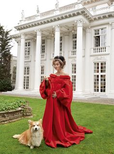 a woman in a red dress standing next to a dog on the grass near a large white building