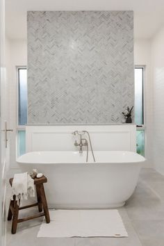 a white bathtub sitting next to a wooden stool in a bathroom under a window