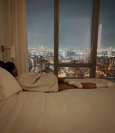 a person laying on a bed in front of a window with the city lights lit up