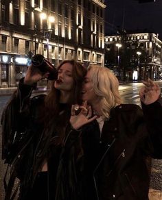 three women standing next to each other on a street corner at night with buildings in the background