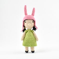 a crocheted doll wearing a green dress and pink bunny ears, standing in front of a white background