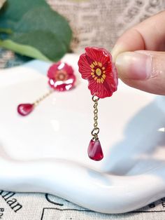 a person is holding a red flower and dangling earrings on a white tray with green leaves