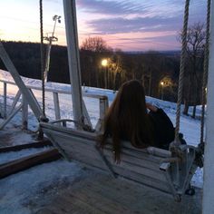 a woman is sitting on a swing in the snow at dusk with her back to the camera