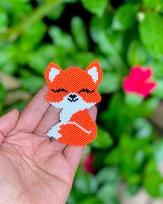 a hand holding up a small red fox brooch in front of green plants and flowers