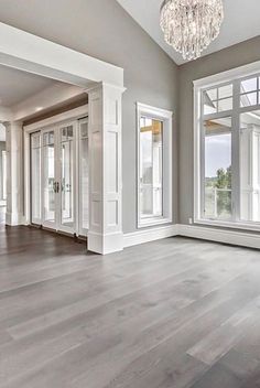 an empty living room with wood floors and chandelier hanging from the ceiling in front of two large windows