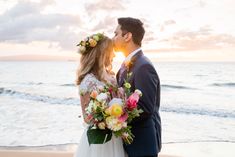 a bride and groom standing on the beach at sunset with their wedding bouquet in front of them