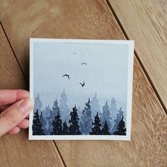 a person holding up a card with trees and birds in the sky behind them on a wooden table