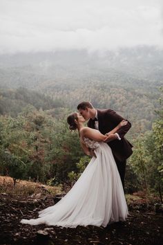 a bride and groom kissing on top of a hill in the mountains with trees behind them