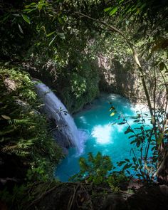 a blue pool in the middle of a forest filled with trees and water flowing down it's sides