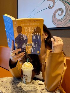 a woman sitting at a table reading a book and drinking milkshake in front of her