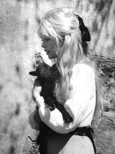 a woman holding a small black animal in her hands while standing next to a stone wall