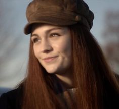 a woman with long red hair wearing a brown hat and smiling at the camera while standing outside