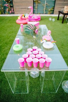 a clear table topped with lots of cups and pink candles on top of green grass