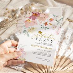 a person holding up a white and pink floral party card with gold foil lettering on it