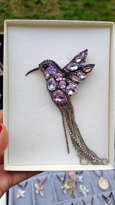 a bird brooch in a white box with purple and silver beads on it's wings