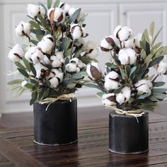 two black vases filled with cotton and greenery on top of a wooden table