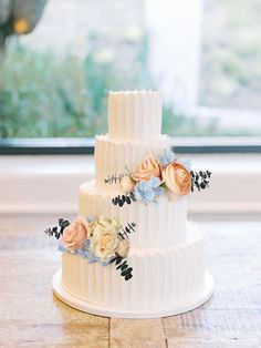 a three tiered white wedding cake with flowers on the top and bottom, sitting on a table in front of a window