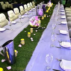 a long table set up with purple and yellow flowers on the grass for a formal function