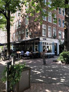 people sitting at tables in front of a tall brick building with many windows and lots of greenery