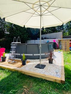 an above ground swimming pool surrounded by gravel and plants with umbrella over it, in the backyard