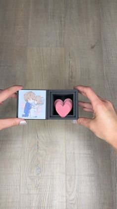 two hands holding an open box with a heart in it on top of a wooden table