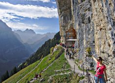 a woman standing on the side of a cliff with her arms in the air and people walking down it