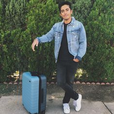 a man standing next to a blue suitcase in front of some bushes and trees with his hand on the handle