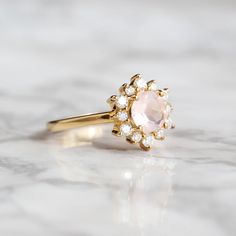 Sometimes when a girl says she likes to receive flowers as a gift, it's not the real flowers she means. Think twice and go for the safest thing - our Flora ring with Rose Quartz and Moissanites. Floral jewelry is where it's at! Perfect as a graduation gift, birthday gift, anniversary ring or promise ring. Details: Center stone Gemstone: Rose Quartz Stone Shape: Round Measurements: approx. 6mm Side stones Gemstone: Moissanite Shape: Round Measurements: 2mm (6), 1.5mm (6) Pictured in vermeil, avai Rose Gold Flower Ring With Center Stone For Promise, Rose Gold Flower Ring With Halo Setting For Promise, Flower Ring With Rose Cut Diamonds For Gifts, Rose Diamond Rings For Anniversary, Rose Gold Crystal Ring For Proposal, Feminine Pink Wedding Rings, Delicate Morganite Rings With Rose Cut Diamonds, Round Cluster Ring With Rose Cut Diamonds For Proposal, Proposal Cluster Ring With Rose Cut Diamonds