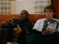 two young men sitting on a couch looking at something in the distance with one man holding his hand out