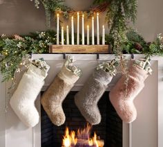 three stockings hanging from a mantel over a fireplace with lit candles and greenery