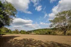 a dirt field with trees and a barn in the distance under a cloudy blue sky