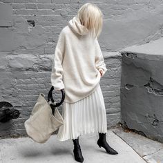 a woman in a white sweater and pleated skirt holding a handbag