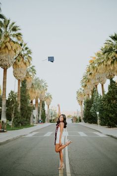 a woman standing in the middle of an empty street with palm trees and a kite flying overhead