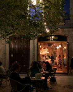 people sitting at tables in front of a building with lights hanging from it's windows