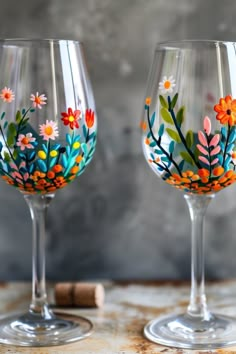 Two wine glasses with floral patterns painted on them, sitting on a wooden surface with a cork nearby. Diy Glassware Projects, Wine Glass Design Ideas, How To Paint On Wine Glasses, Painting Ideas On Glass Cups, Art And Wine Painting Ideas, Acrylic Wine Glass Painting, Floral Wine Glass Painting, Hand Painted Glasses Diy, Painting On Wine Glasses Diy