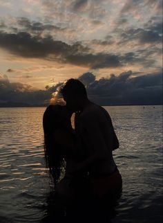 a man and woman are kissing in the water at sunset with clouds above their heads