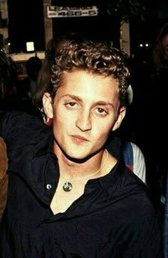 a young man with curly hair wearing a black shirt and looking at the camera while standing in front of a crowd