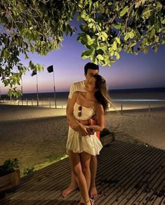 a man and woman standing under a tree on the beach at night with their arms around each other