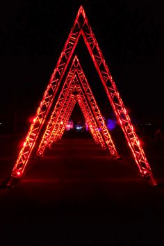 Very large, red triangles in a row. Triangles are made out of metal trussing with red light inside each piece of trussing. Triangle structures our outside at night. Large enough to walk through. Festival Stage Design Outdoor, Event Stage Design Ideas, Event Tunnel Entrance, Outdoor Stage Design, Truss Decor, Coachella Decor, Neon Entrance Arch, Outdoor Event Lighting