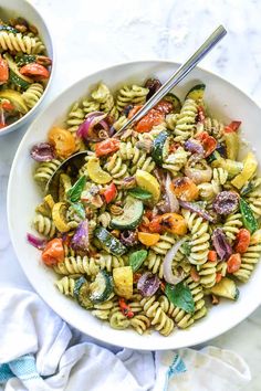 two bowls filled with pasta salad on top of a white tablecloth next to silver spoons