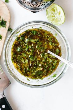 a glass bowl filled with green salsa next to a cutting board and lime wedges