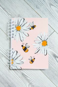 a pink notebook with white daisies and bees on it, sitting on a wooden surface