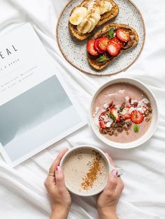 a person holding a cup of coffee next to two plates with food on them and an open book