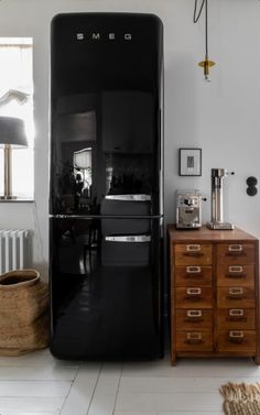 a black refrigerator sitting in the middle of a room next to a dresser and lamp