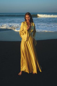 a woman in a long yellow dress standing on the beach with her hand up to her chest